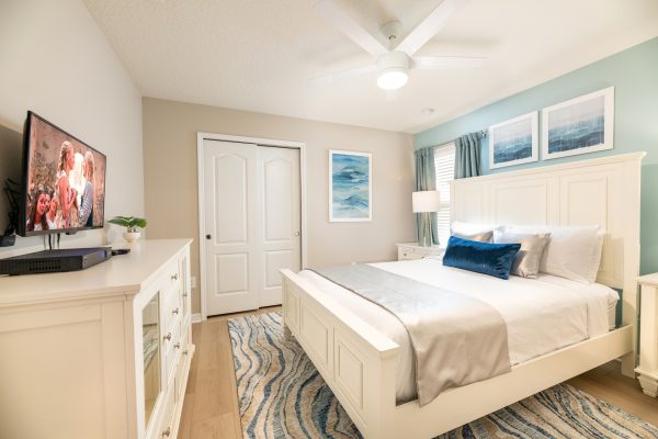 Coastal white bedroom with flat-screen TV on dresser and blue accent rug at Magic Moments Kissimmee vacation villa